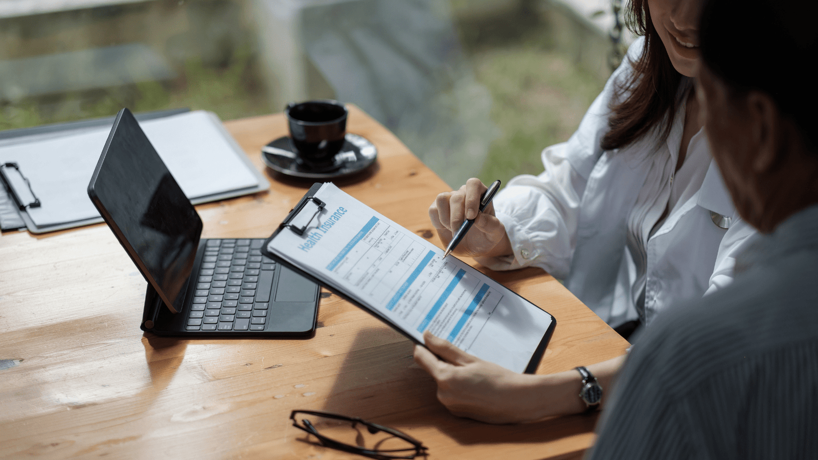 Une personne qui discute d'une assurance maladie avec un médecin ou un conseiller et qui remplit un formulaire intitulé « Health Insurance ». Sur la table se trouvent un ordinateur portable et une tasse de café.