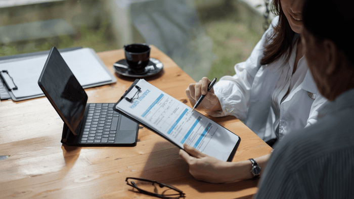 Une personne qui discute d'une assurance maladie avec un médecin ou un conseiller et qui remplit un formulaire intitulé « Health Insurance ». Sur la table se trouvent un ordinateur portable et une tasse de café.