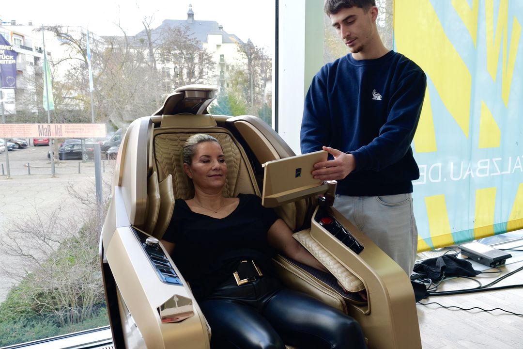 Eine Kundin sitzt zurückgelehnt in einem Massagesessel, ein Mitarbeiter zeigt ihr die Einstellungen am Display
